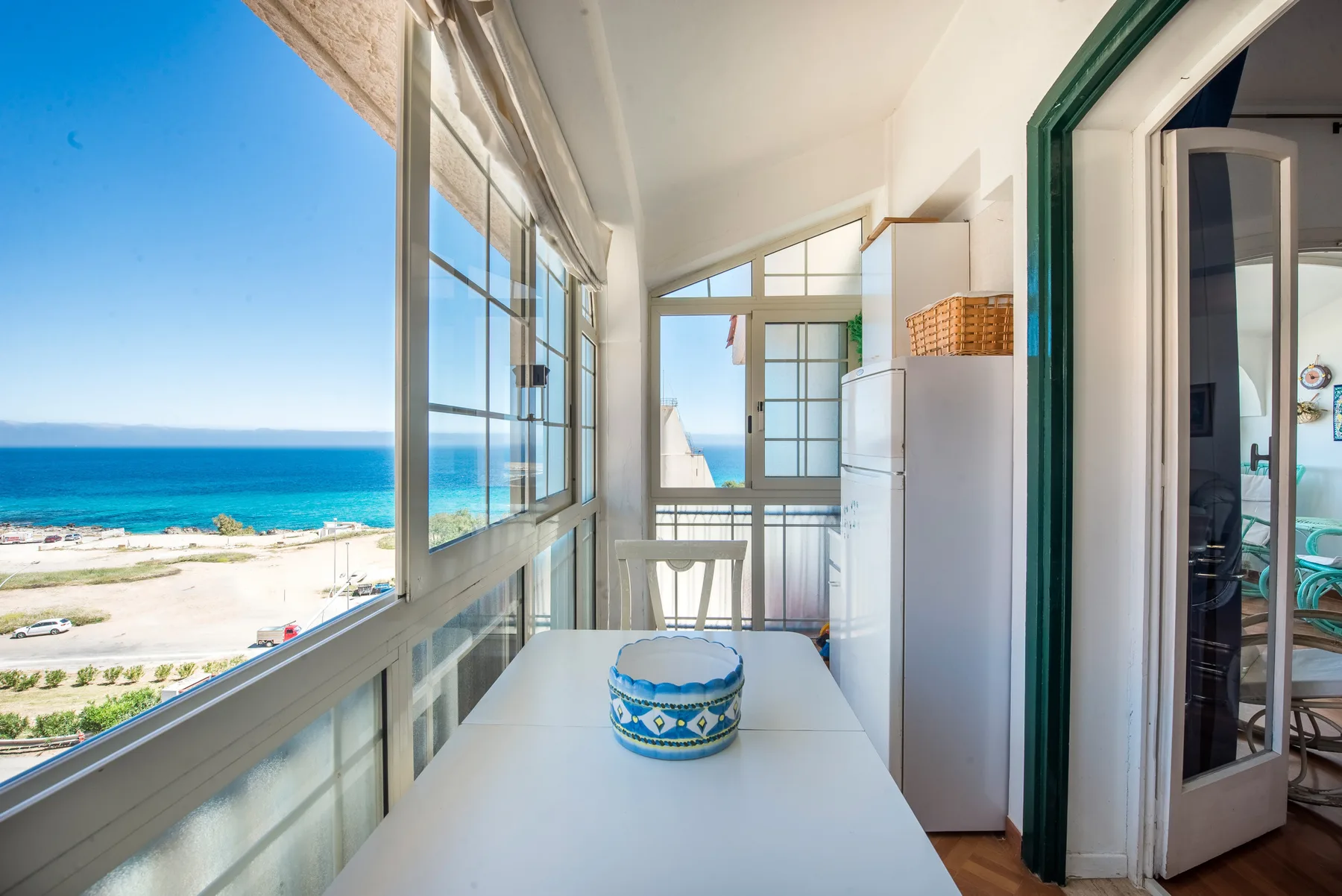 A bright enclosed balcony with a table and windows overlooking the sea.