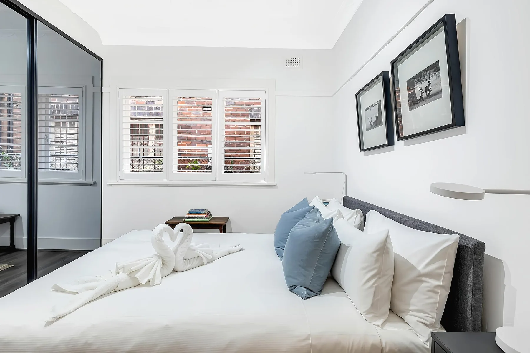Un dormitorio luminoso con una cama bien hecha y ventanas con persianas blancas.
