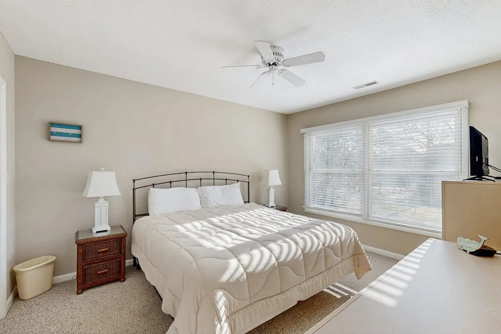 A bedroom with a bed, bedside tables, and a large window with blinds.