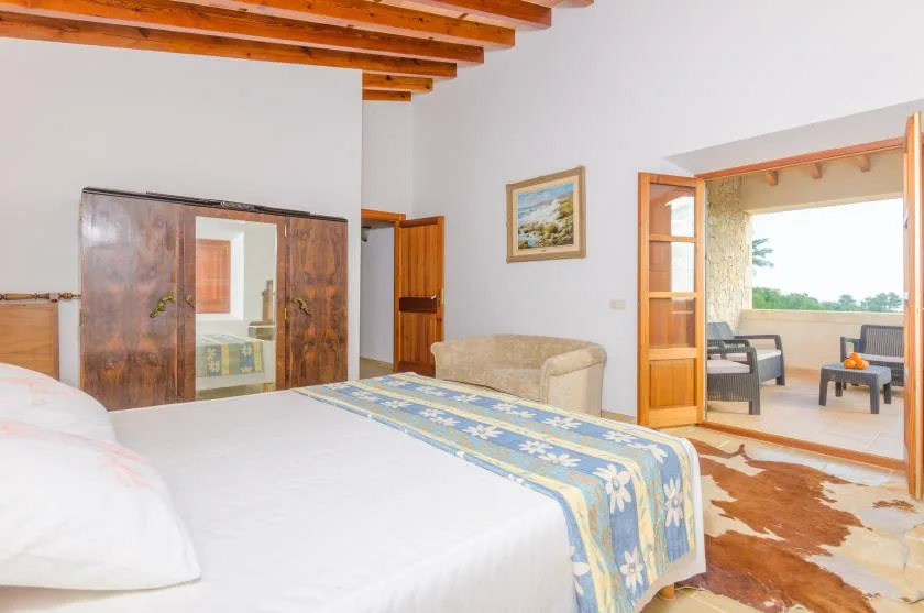 Un dormitorio con una cama, una zona de asientos beige y una puerta abierta a una sala luminosa.