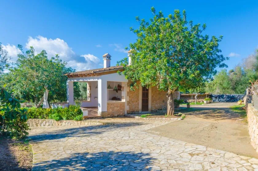 Un edificio bajo de piedra y yeso junto a un patio pavimentado y un árbol frondoso.