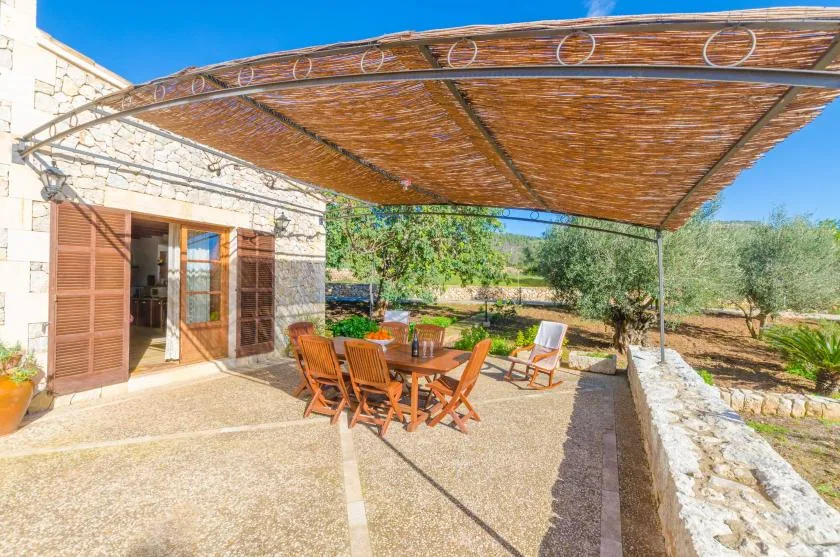Un patio comedor a la sombra junto a una casa de piedra, con asientos de madera y vegetación.