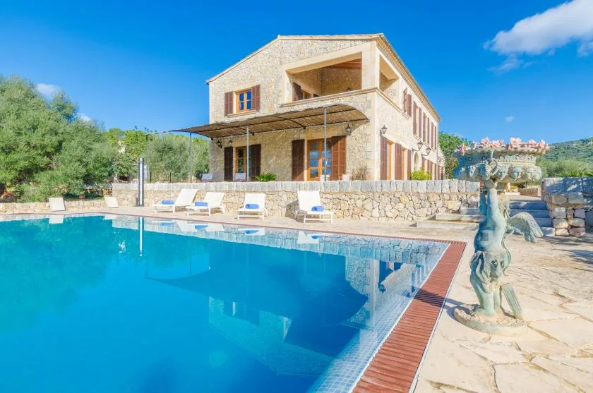 Una casa de piedra está junto a una piscina, con tumbonas en el borde opuesto.