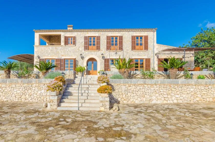 Una casa de piedra con ventanas con contraventanas se alza tras un amplio patio y escalones centrales.