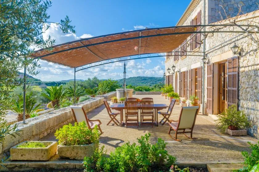 Una terraza comedor a la sombra junto a un edificio de piedra, con vistas a colinas verdes.