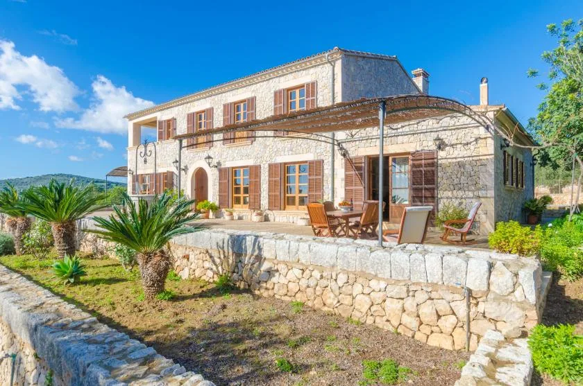 Una casa de piedra con terraza, asientos al aire libre y palmeras en el jardín.