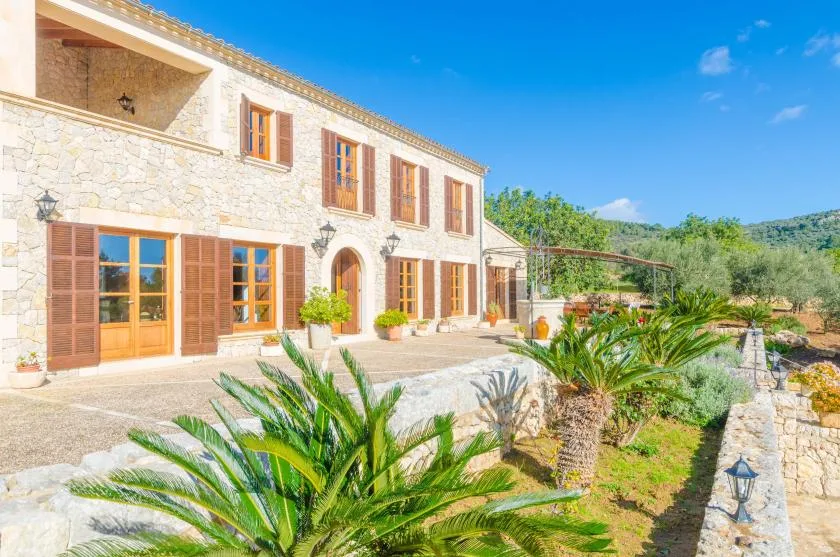 Una casa de piedra con ventanas con contraventanas da a un jardín bajo un cielo despejado.