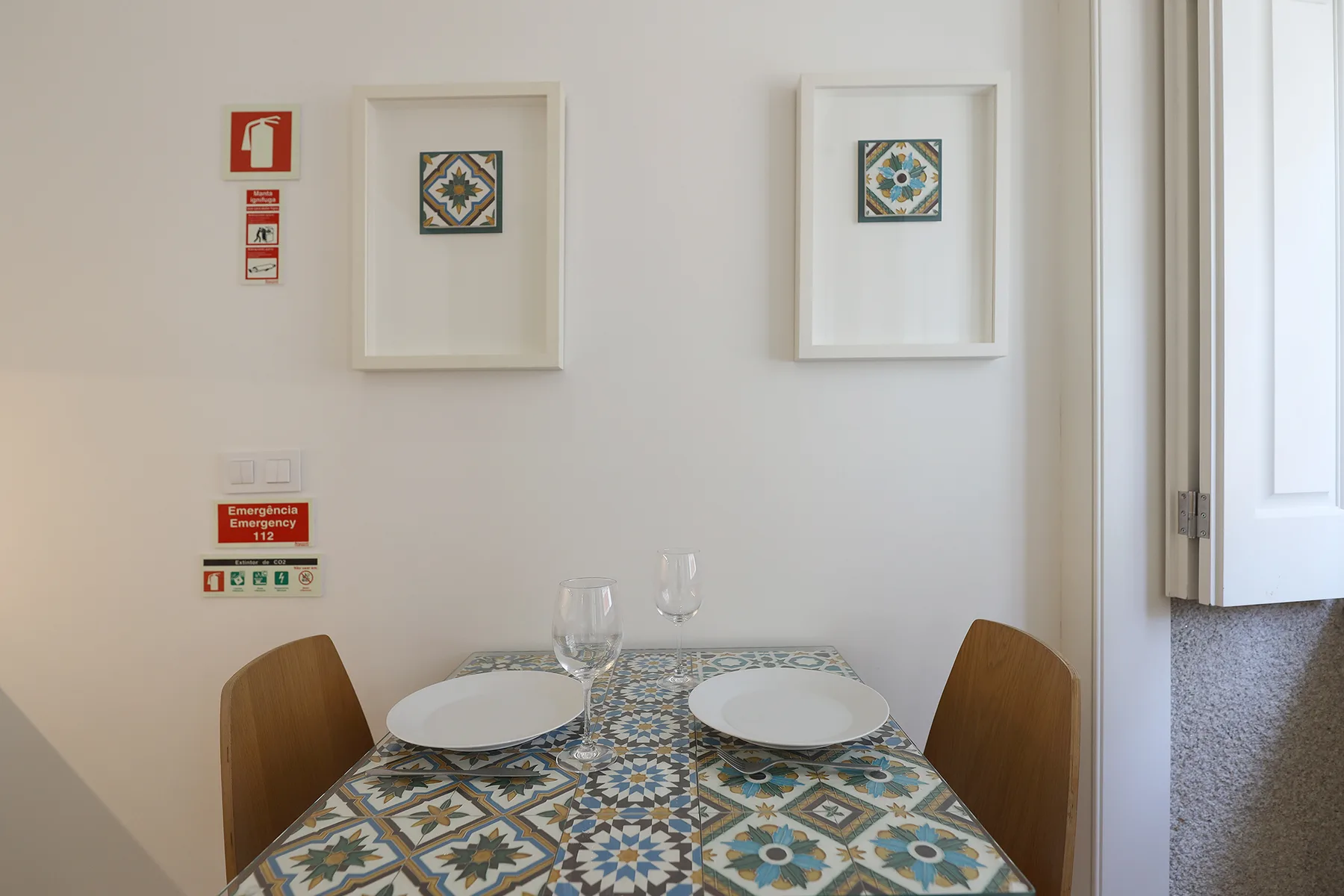 A dining table with patterned tile and two place settings sits beside a bright window.
