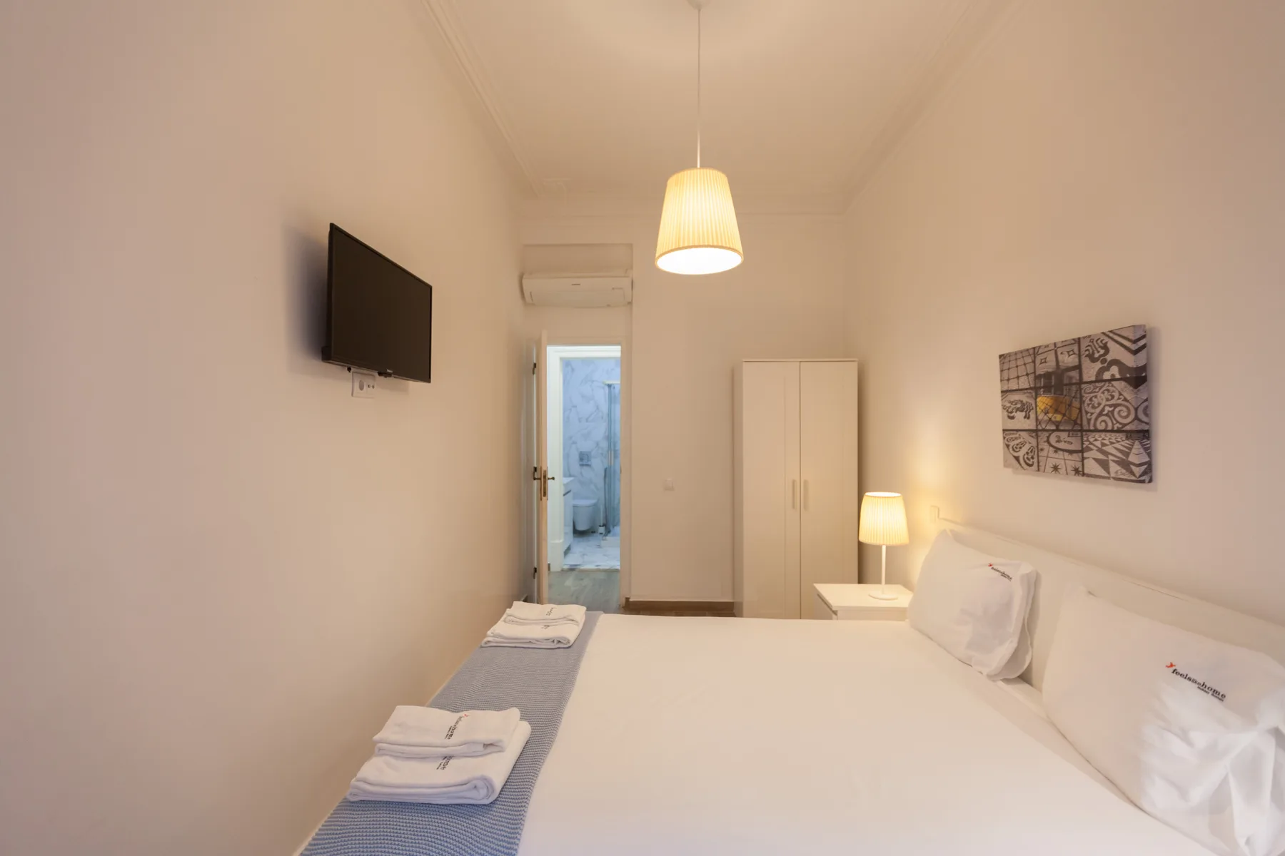Un dormitorio luminoso con ropa de cama blanca, un televisor en la pared y un armario.
