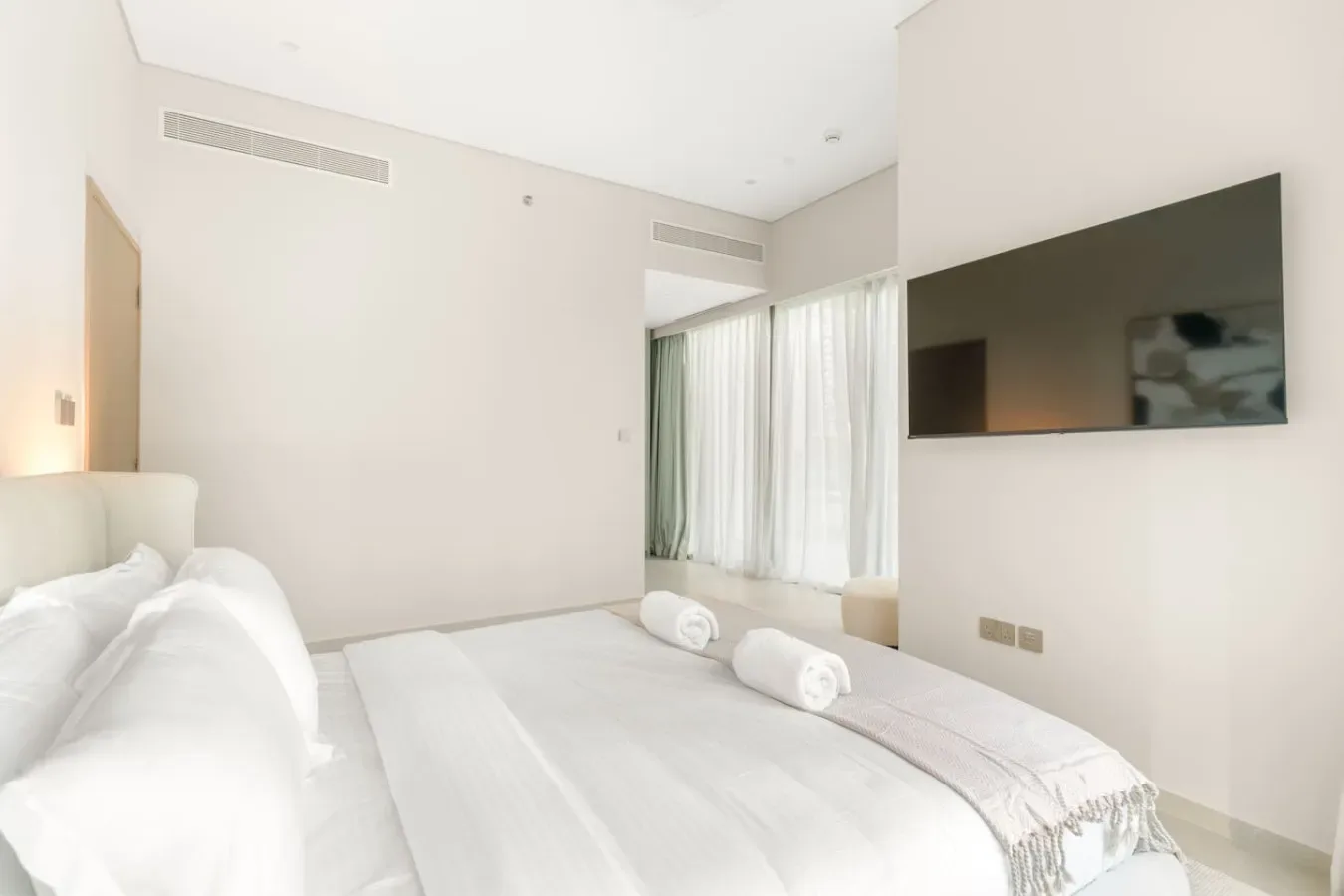 Un dormitorio luminoso con ropa de cama blanca y un televisor en la pared.