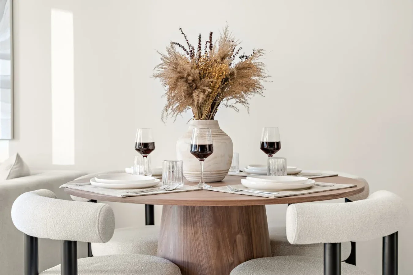 Una mesa de comedor está puesta con platos, cristalería y un jarrón con flores secas.