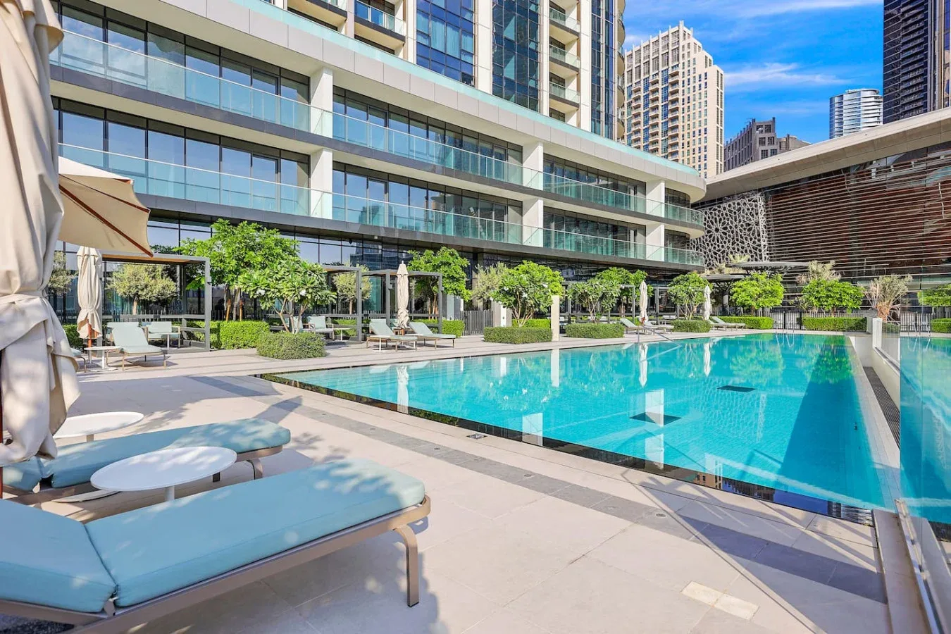 Una piscina al aire libre junto a un edificio moderno de gran altura, con tumbonas cerca.