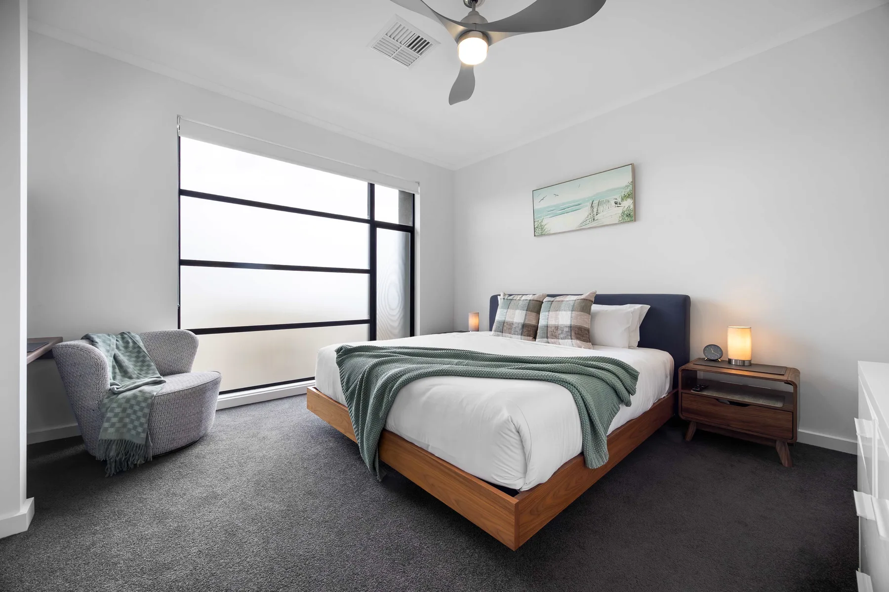 A bedroom with a neatly made bed, an armchair, and a large window.