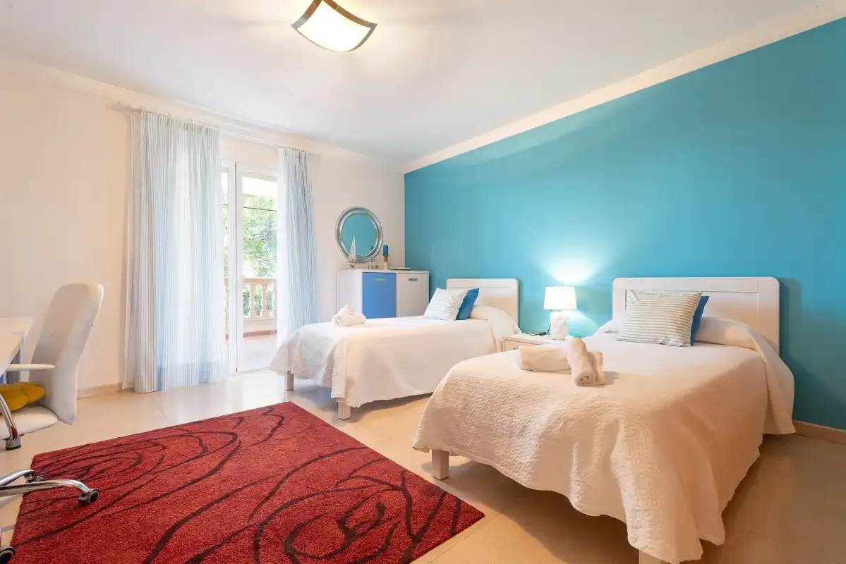 A bedroom with two beds, a turquoise accent wall, and a red rug.