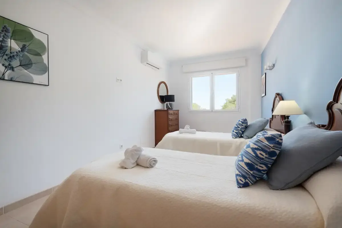 A bright bedroom with two beds, blue accent pillows, and a window.
