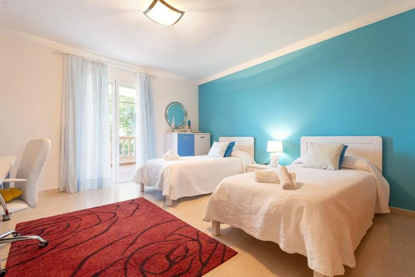 A bright bedroom with two beds, a teal accent wall, and a red rug.