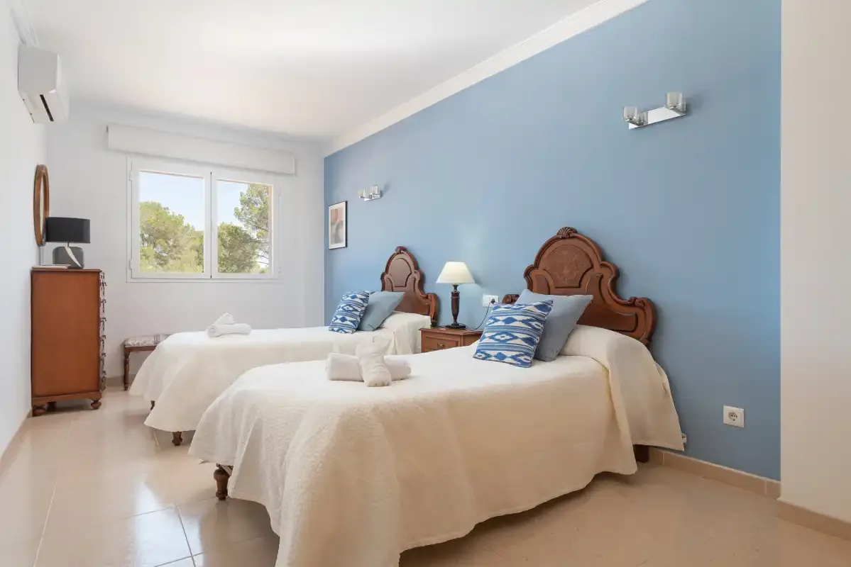 A bright bedroom with two beds, blue walls, and a window overlooking greenery.