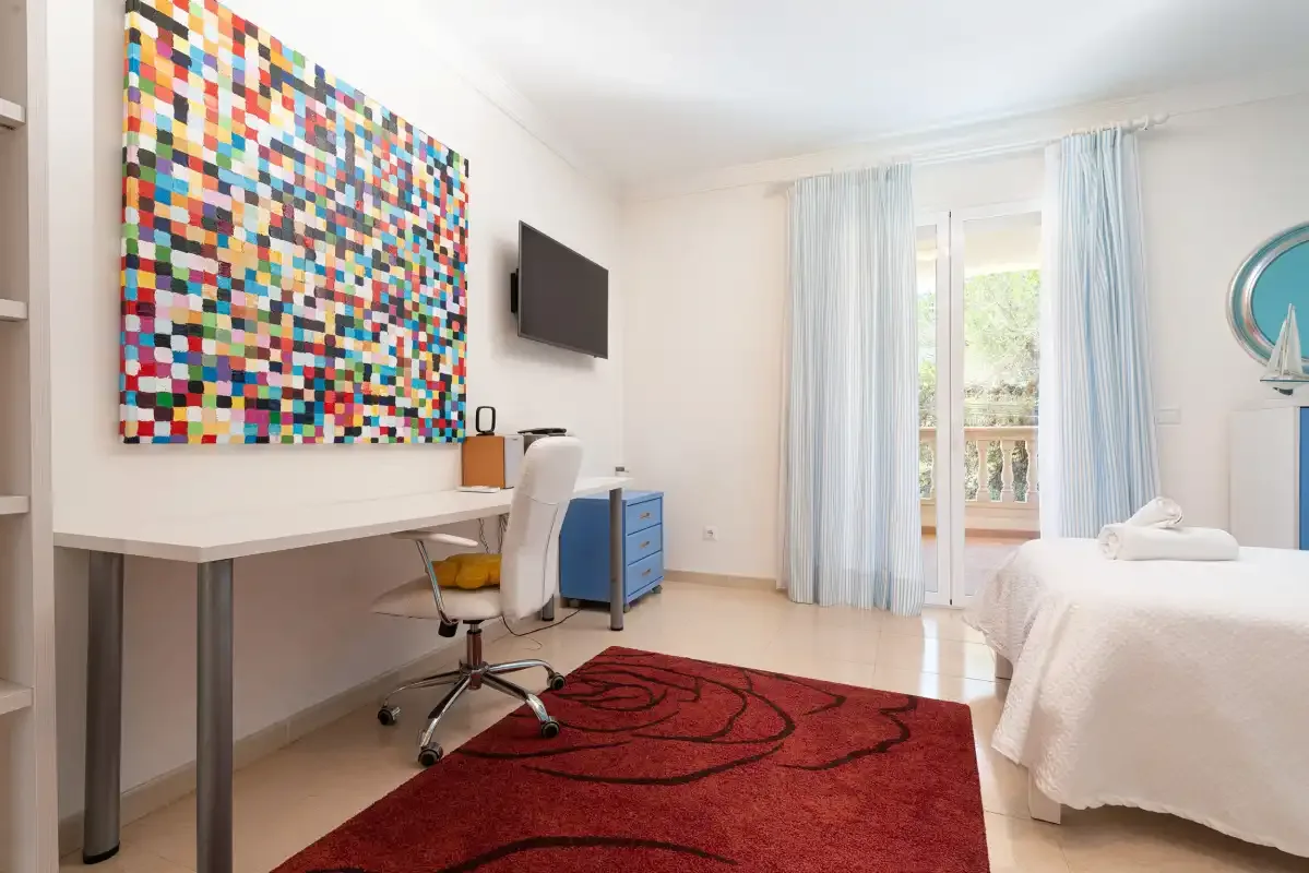 A bright bedroom with a desk, colorful wall art, and a bed beside the window.