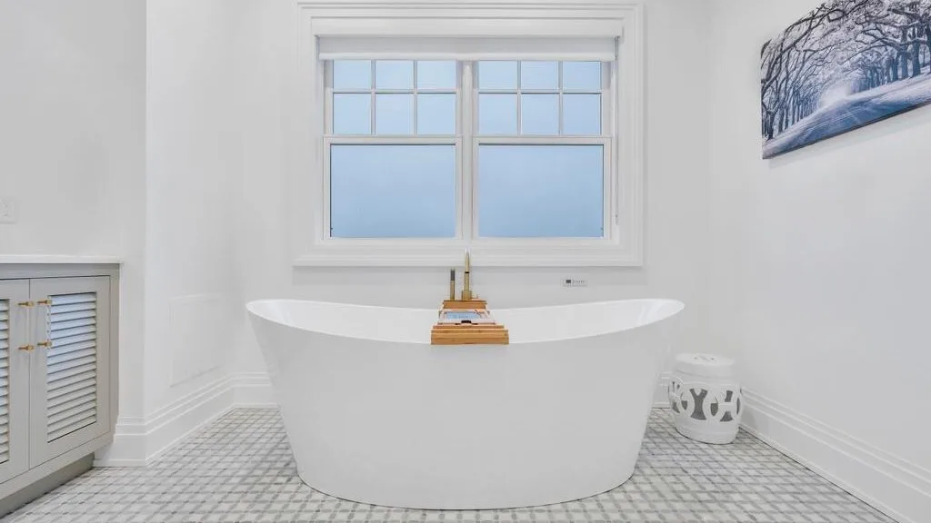 Una bañera independiente está bajo una ventana grande en un baño luminoso.
