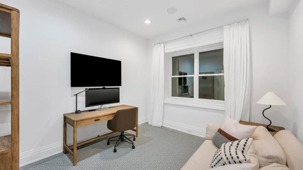 Una sala luminosa con escritorio, televisor y asientos junto a una ventana con cortinas.
