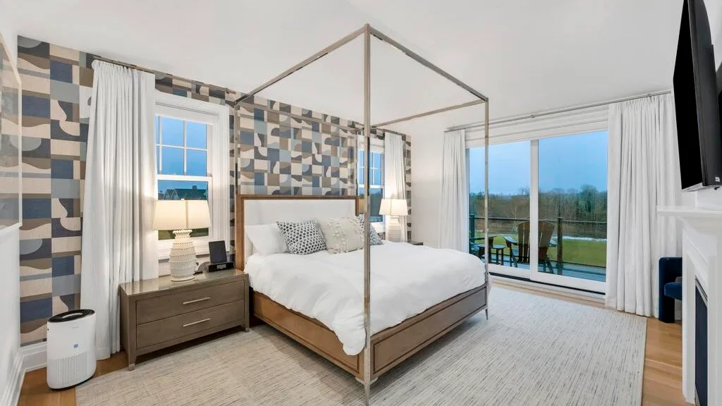 Un dormitorio con una cama con dosel junto a grandes ventanas y paredes estampadas.