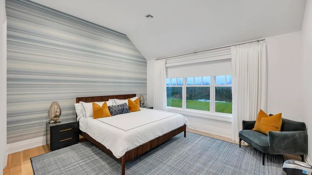Un dormitorio con una cama, una pared decorativa a rayas y asientos grises junto a la ventana.