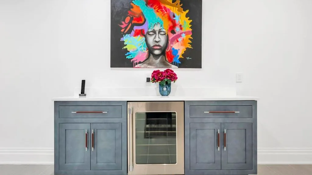 Un mueble empotrado con una nevera de bebidas con puerta de vidrio está bajo un retrato colorido.