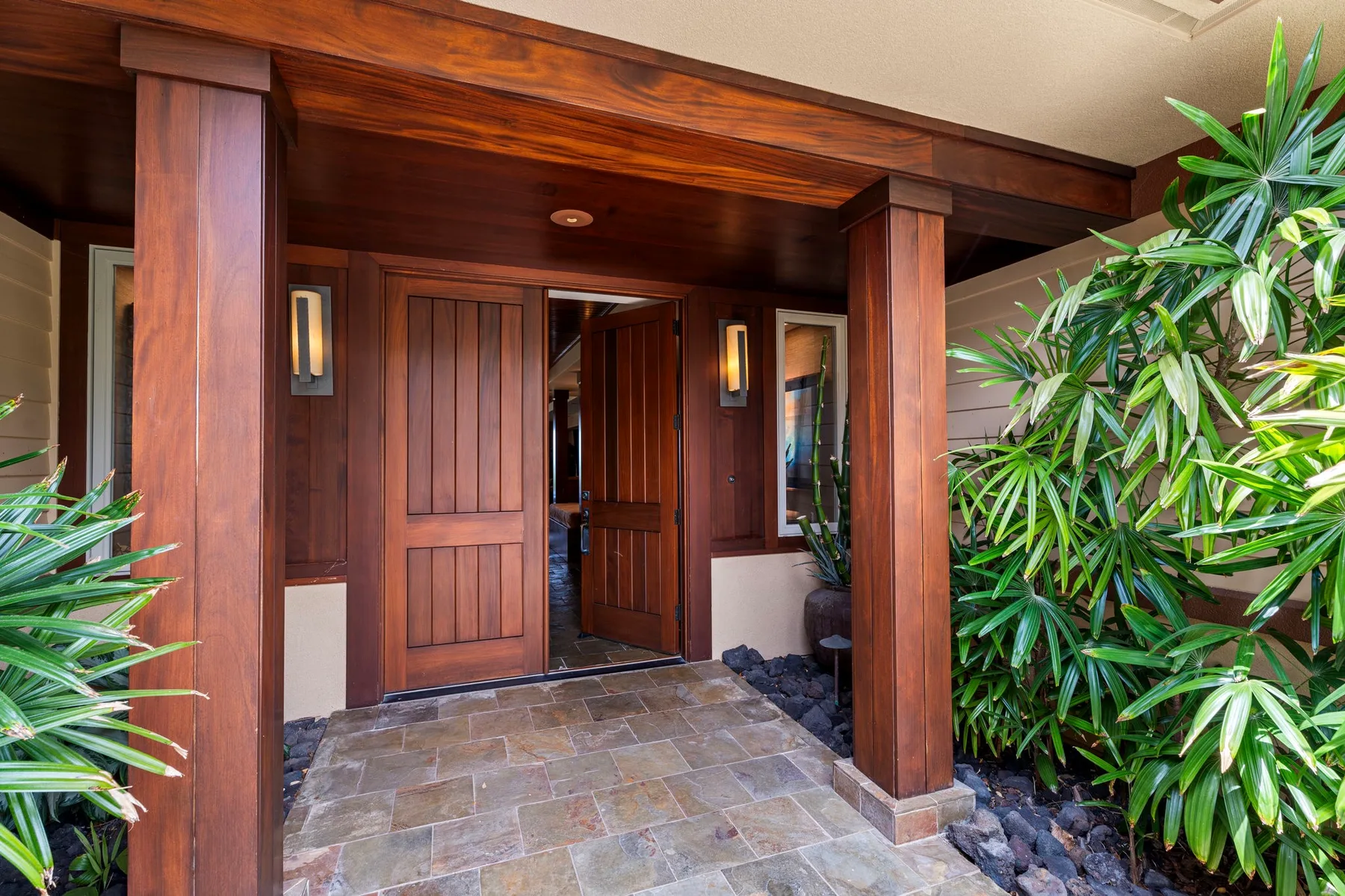 A covered entrance with wooden doors and leafy plants beside a tiled walkway.