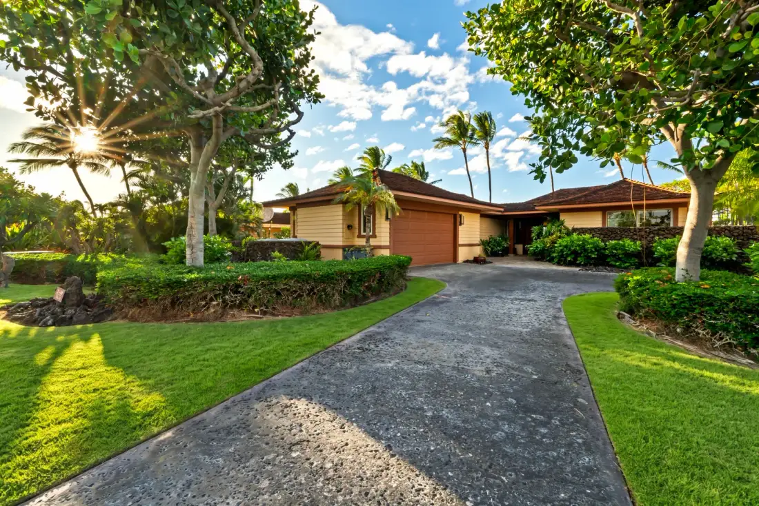A single-story house sits at the end of a paved driveway, surrounded by trees and lawn.