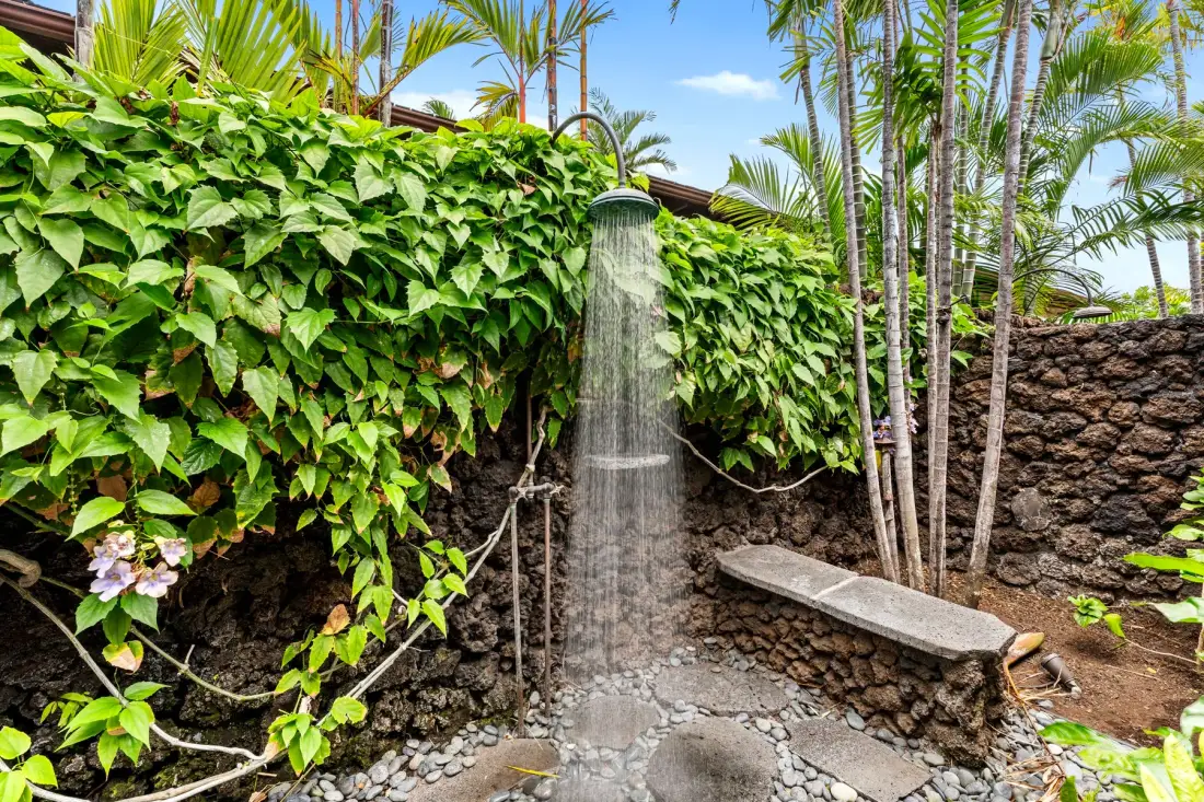 An outdoor shower stands beside a stone wall covered in lush greenery.