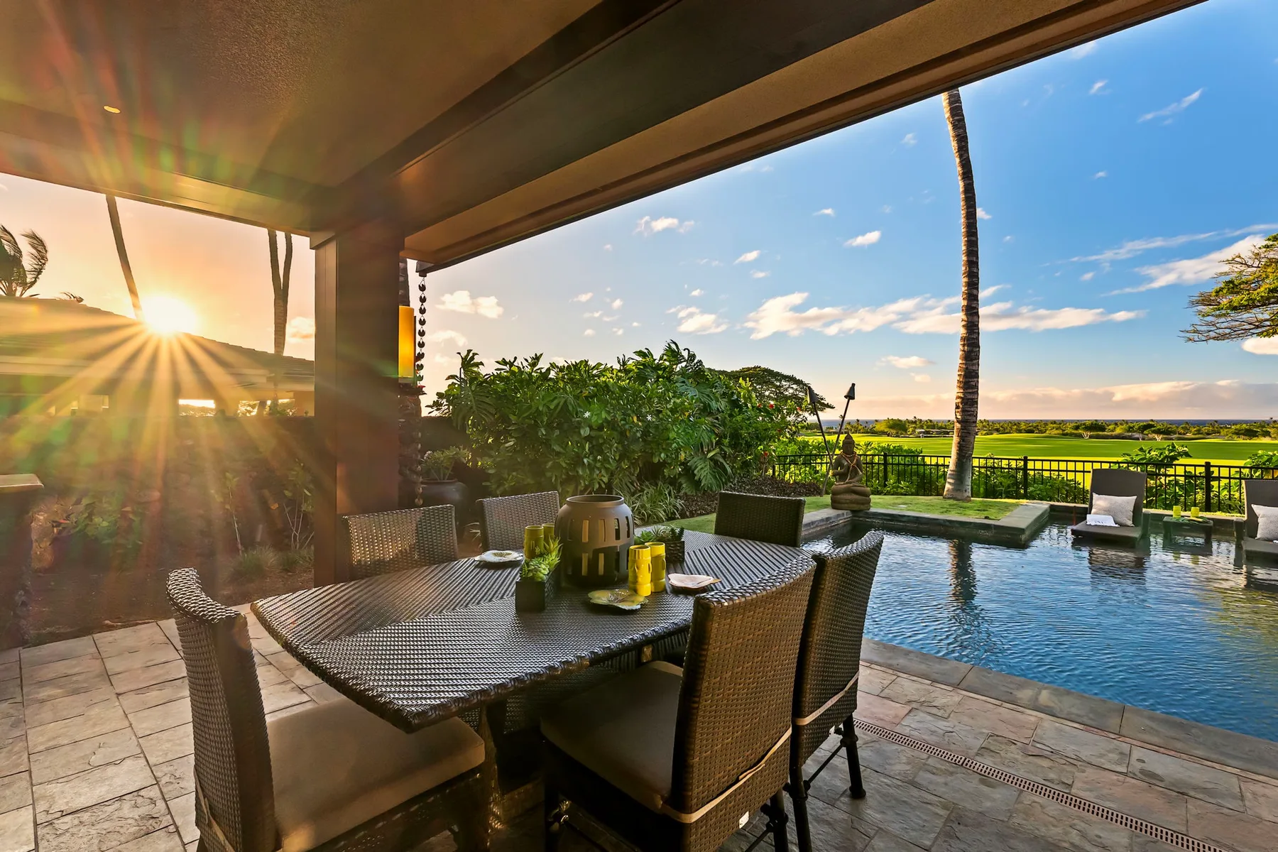 A covered patio has a dining table beside a swimming pool and green landscape.