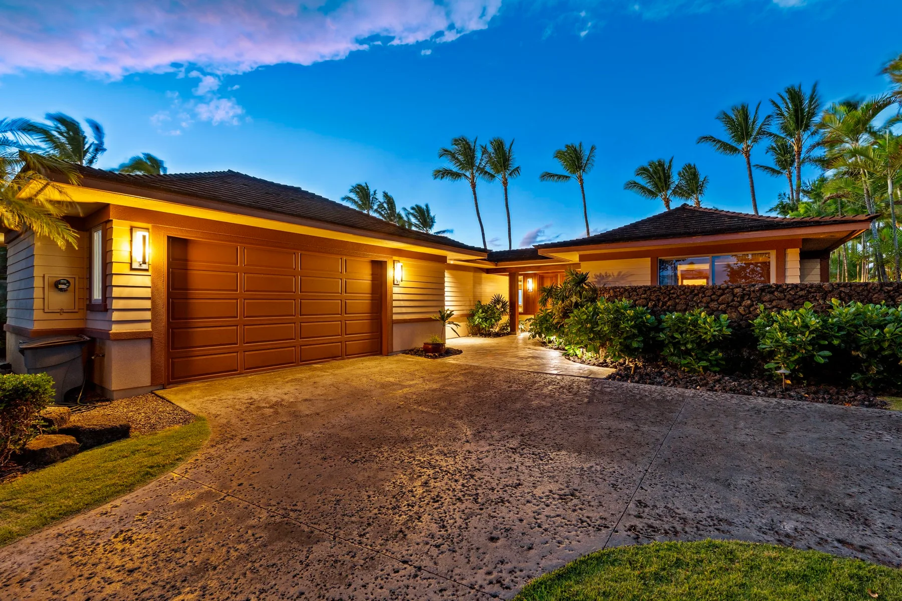 A house exterior at dusk, with a wide driveway and palm trees.