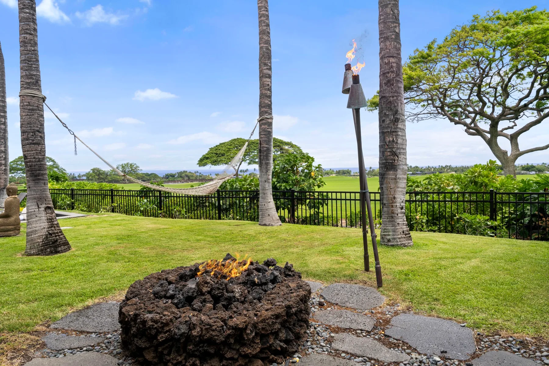 A landscaped garden with a stone fire pit, palm trees, and an ocean view.