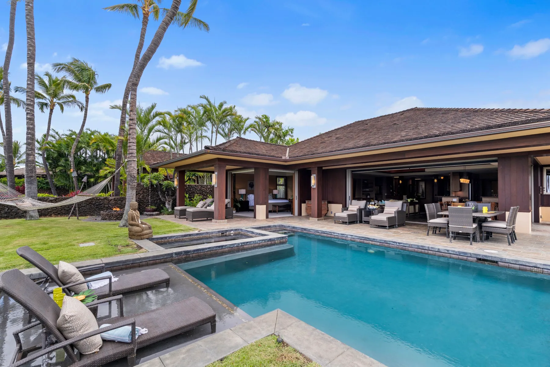 A swimming pool sits beside a covered patio with outdoor seating and palm trees.