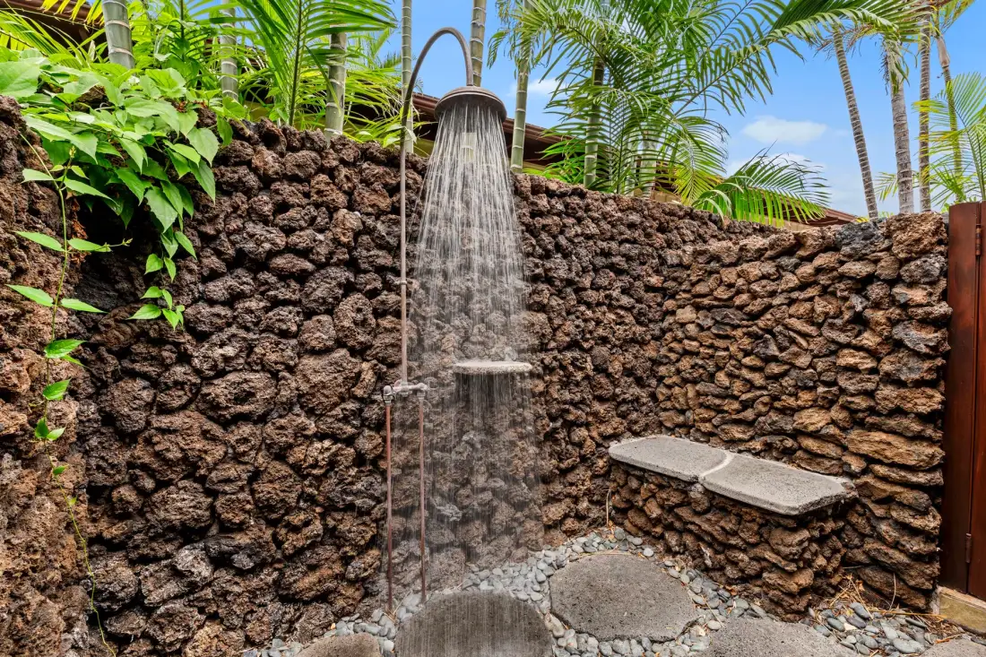 An outdoor shower beneath palm trees, bordered by a rough stone wall.