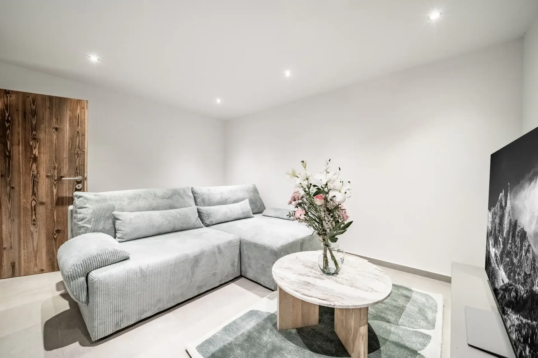 Minimalist living room with a large gray sectional sofa, a vase of flowers on a round table, and recessed lighting illuminating the space.