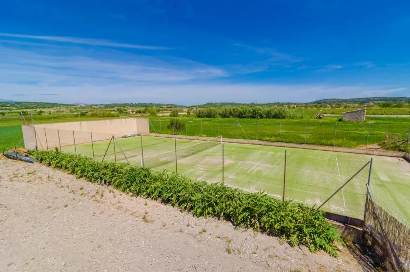 A fenced outdoor tennis court sits beside open green fields.