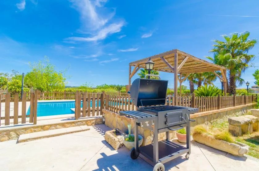 A poolside patio features a barbecue grill beside a wooden pergola.