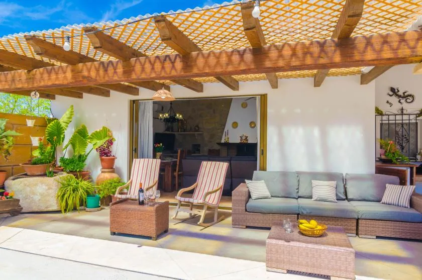 An outdoor patio seating area sits beneath a wooden pergola, with plants nearby.