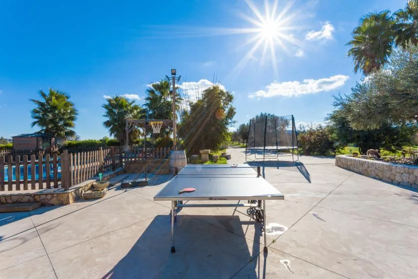 An outdoor recreation area with a ping-pong table and a fenced court under the sun.