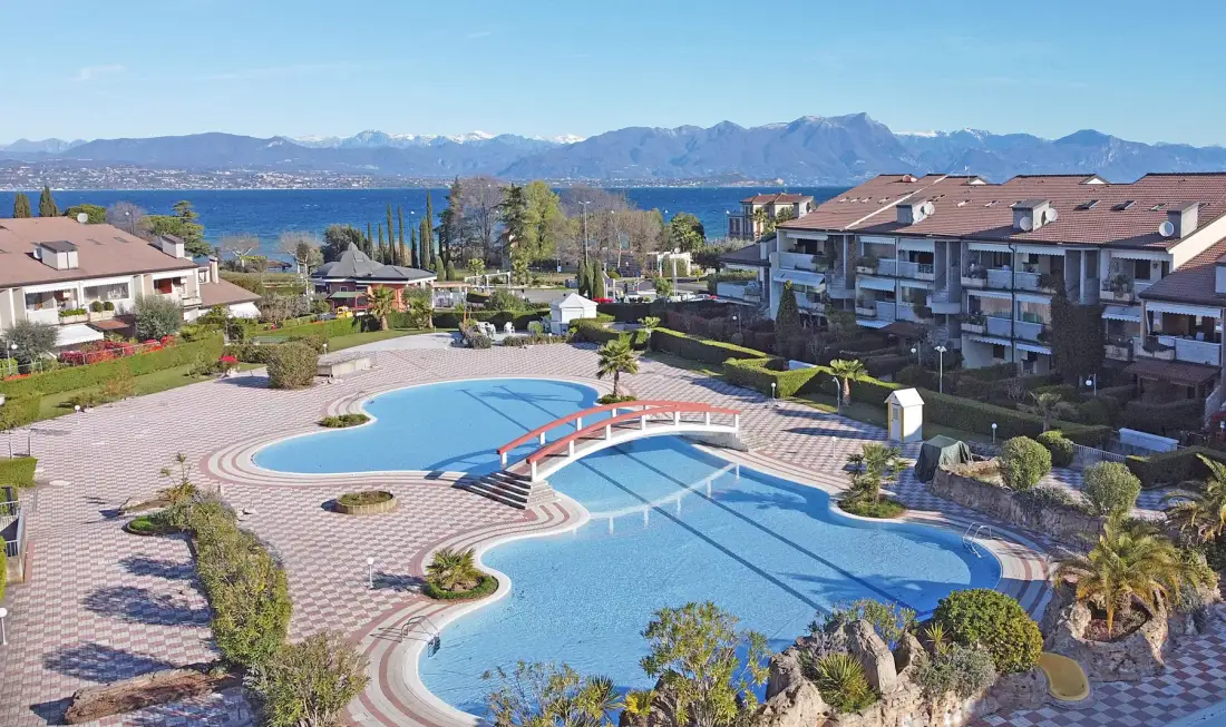 A landscaped outdoor swimming pool sits among residential buildings, with a lake and mountains beyond.
