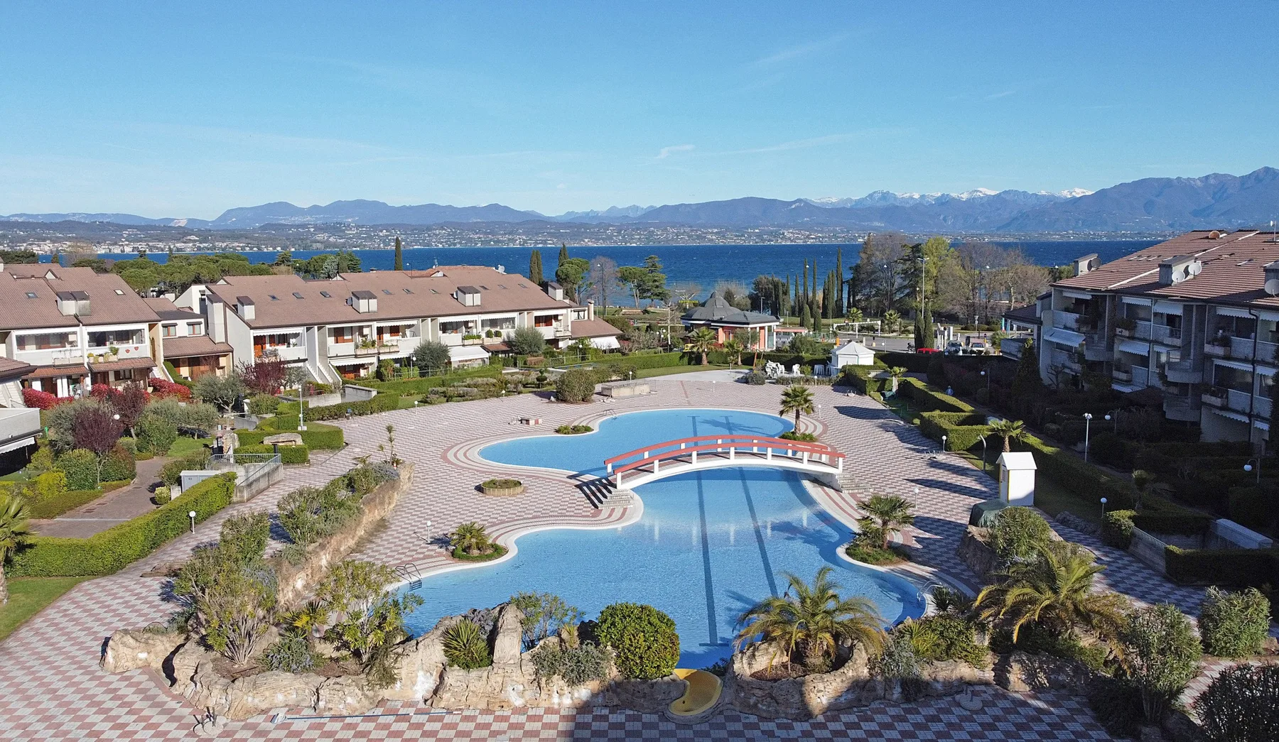 A landscaped outdoor pool sits among residential buildings, with a lake and mountains in the distance.