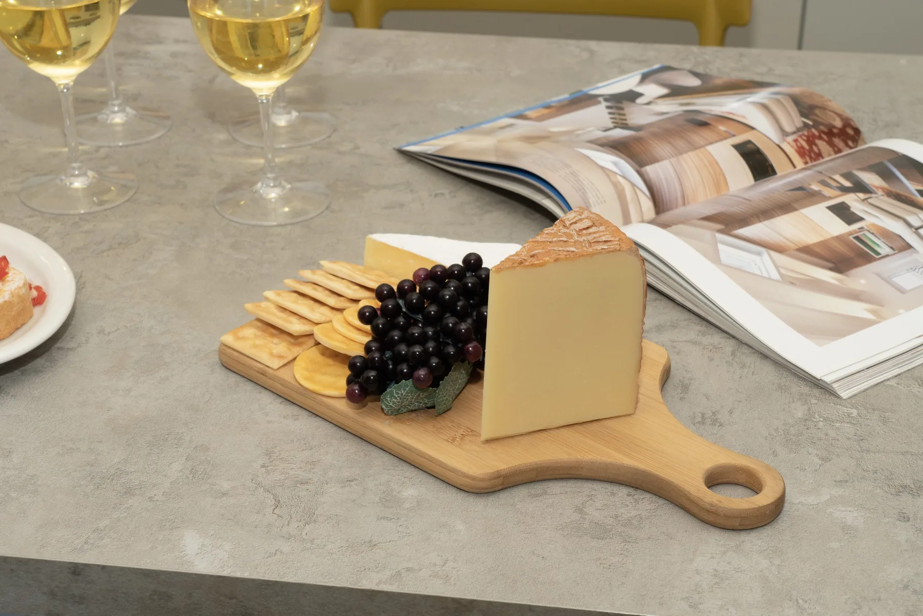 A cheese and fruit board sits beside an open magazine and wine glasses.