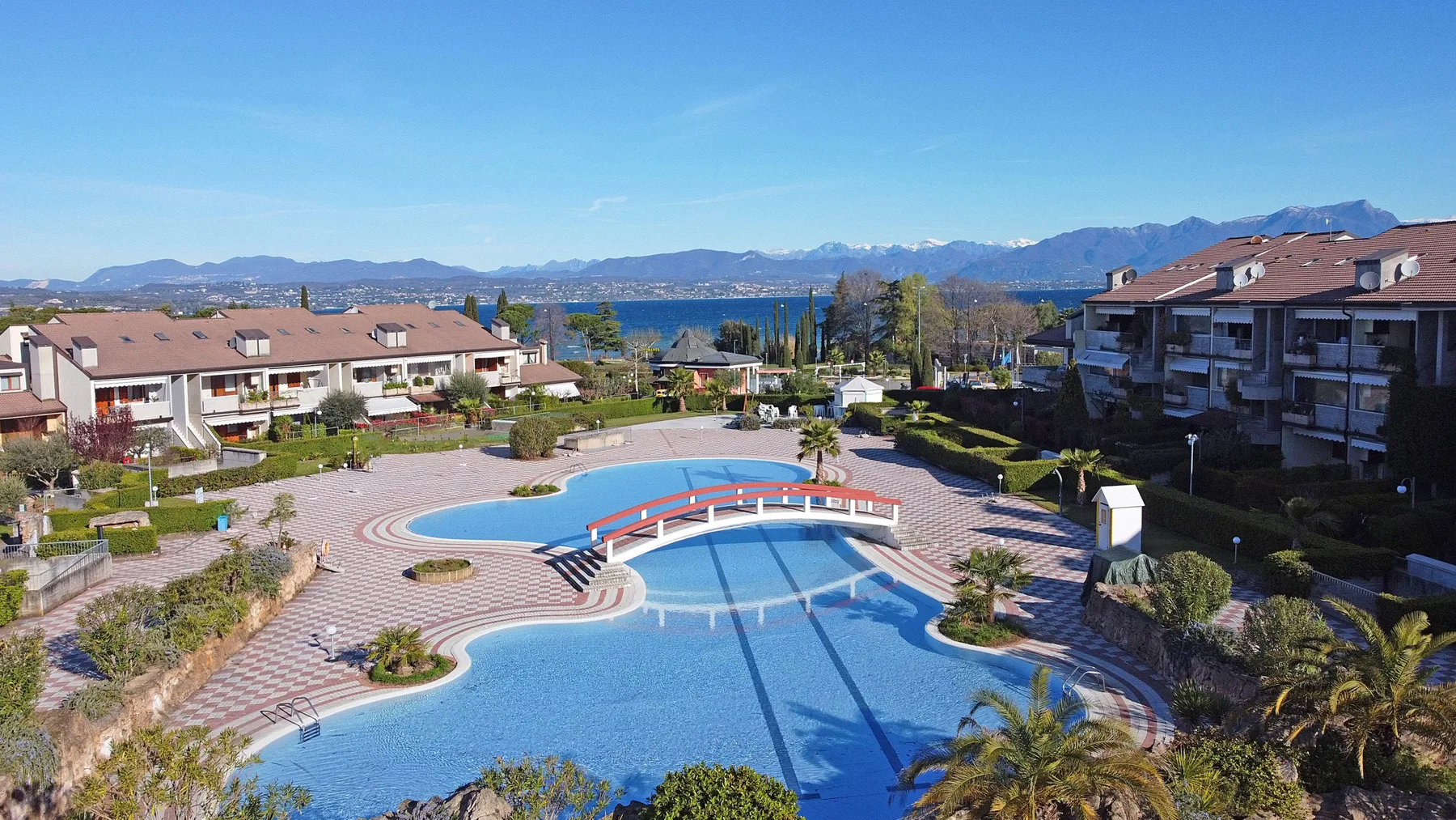 A curved outdoor pool sits among residential buildings, with a lake and mountains beyond.