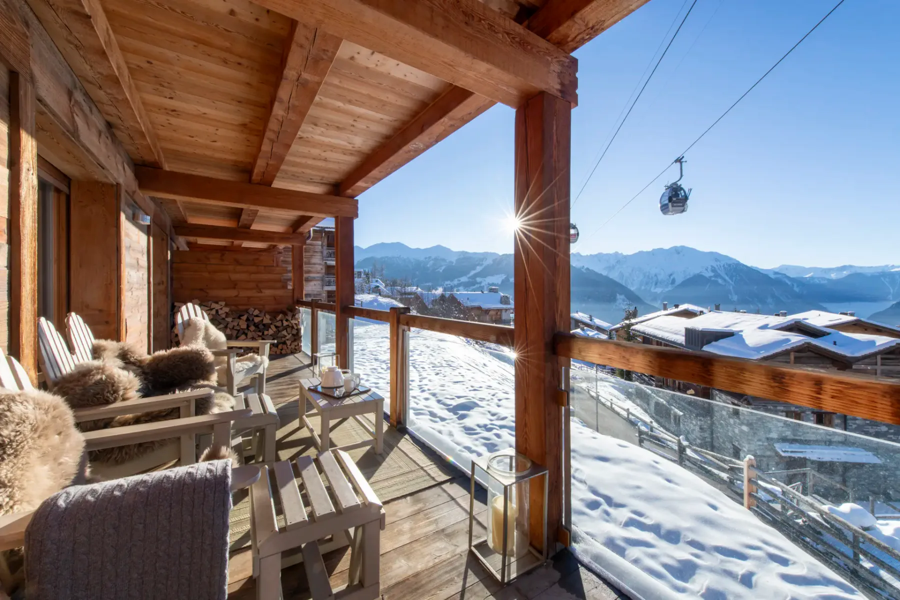 A wooden terrace with chairs and potted plants overlooks snowy rooftops and mountain slopes; a ski gondola is visible in the background.
