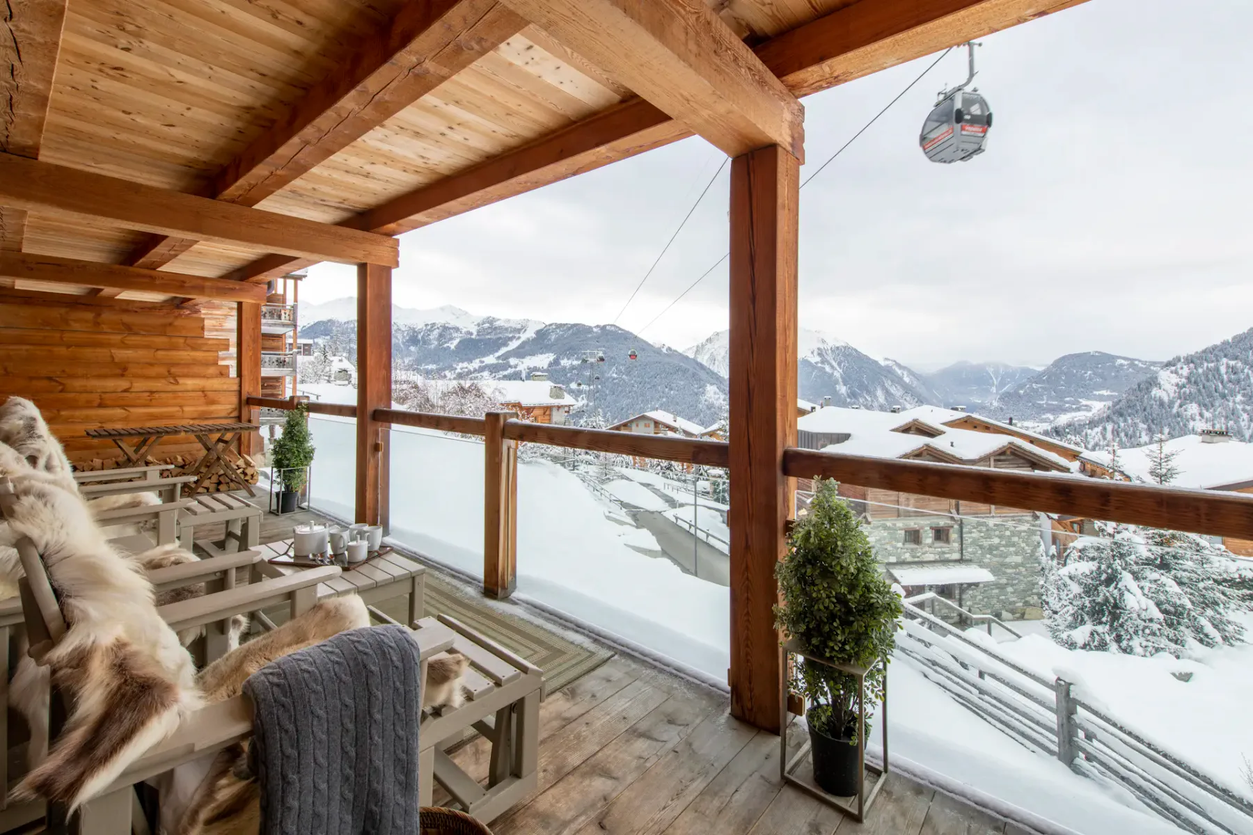 A wooden terrace with chairs and potted plants overlooks snowy rooftops and mountain slopes; a ski gondola is visible in the background.