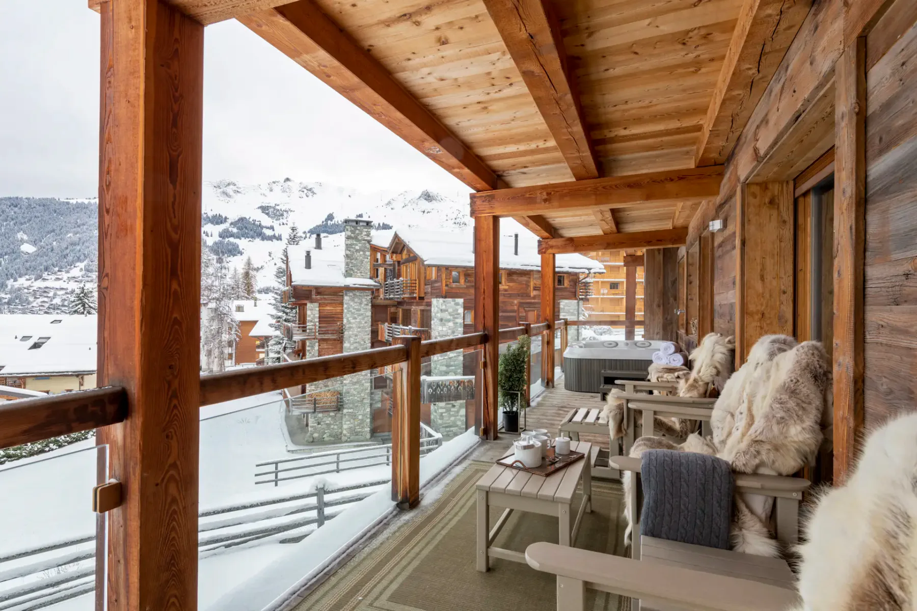 A wooden terrace with chairs and potted plants overlooks snowy rooftops and mountain slopes; a ski gondola is visible in the background.