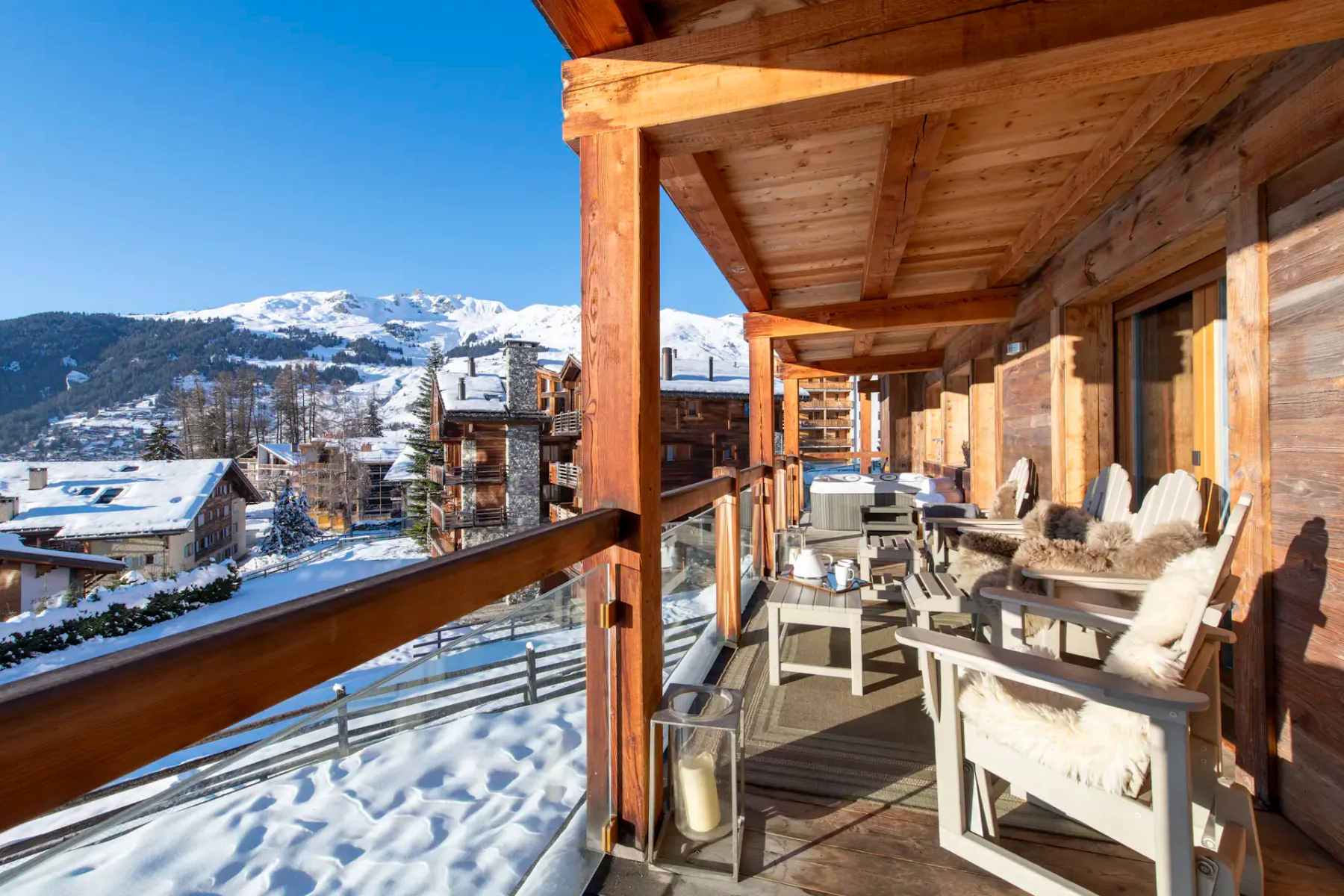 A wooden terrace with chairs and potted plants overlooks snowy rooftops and mountain slopes; a ski gondola is visible in the background.