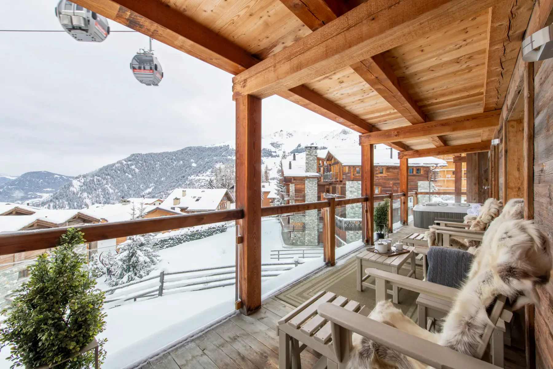 A wooden terrace with chairs and potted plants overlooks snowy rooftops and mountain slopes; a ski gondola is visible in the background.