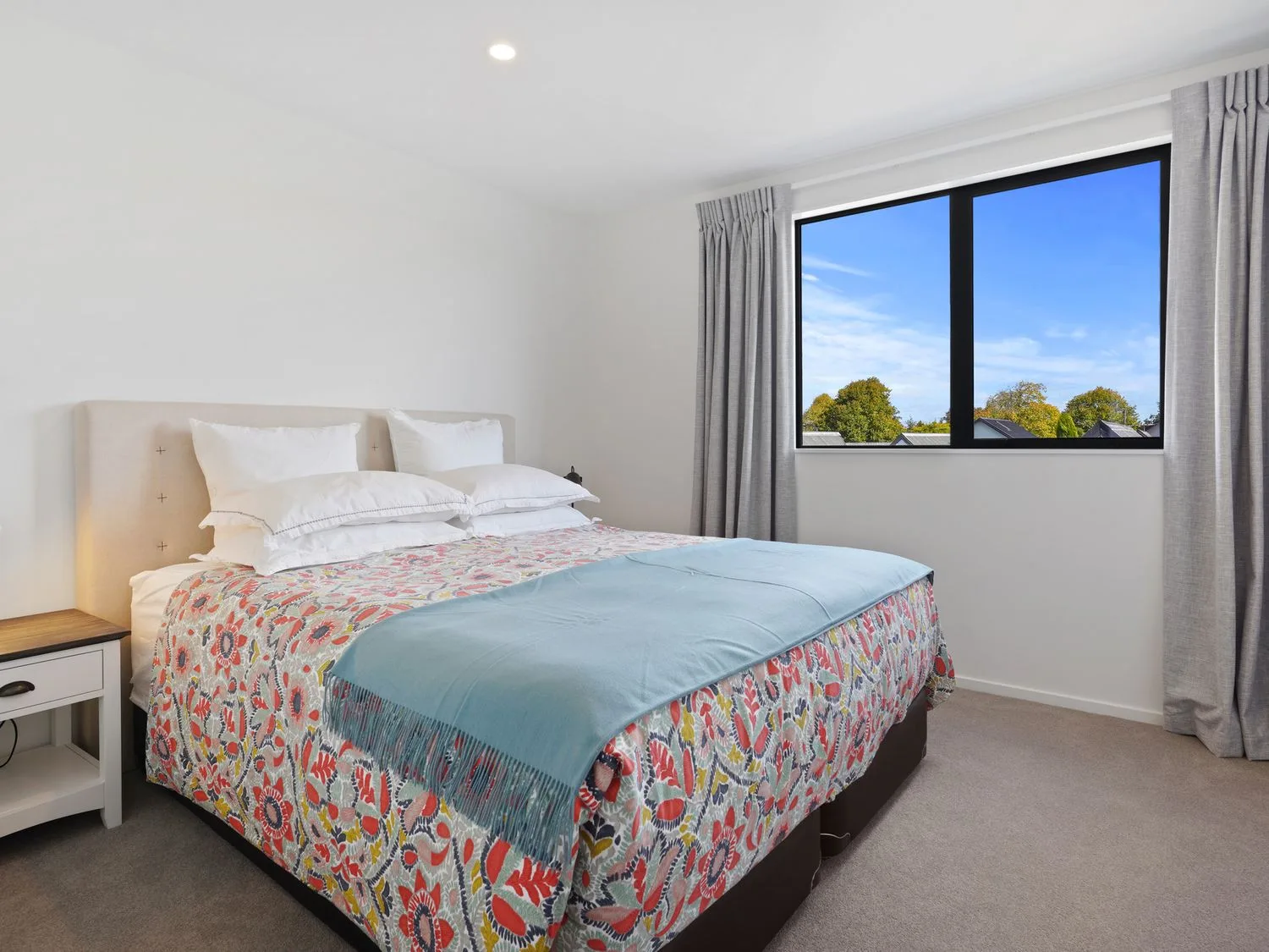 Un dormitorio con una colcha floral y una manta azul junto a una ventana con cortinas grises.